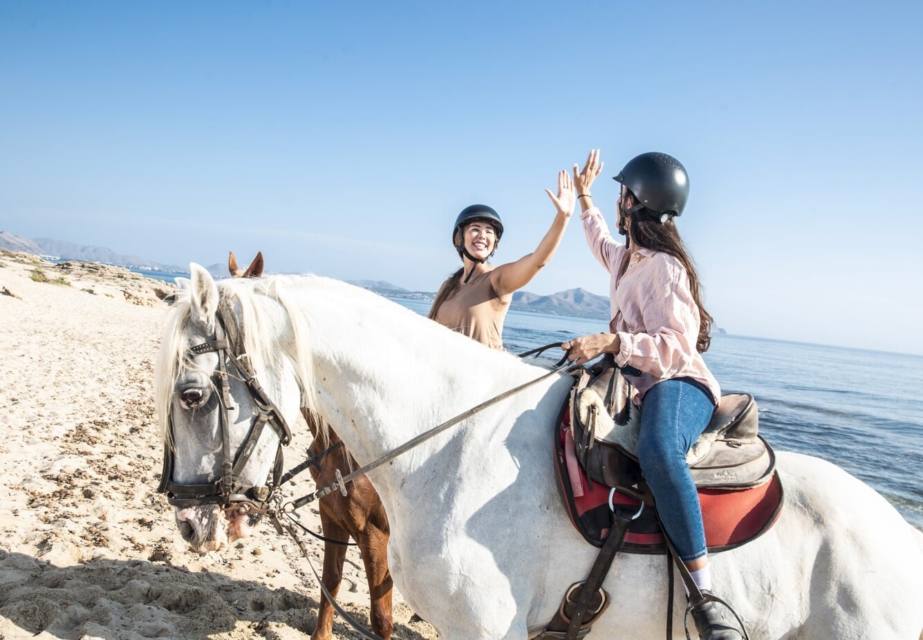 Friends riding horses on the beach on a sunny day in Mallorca celebrating together by the sea