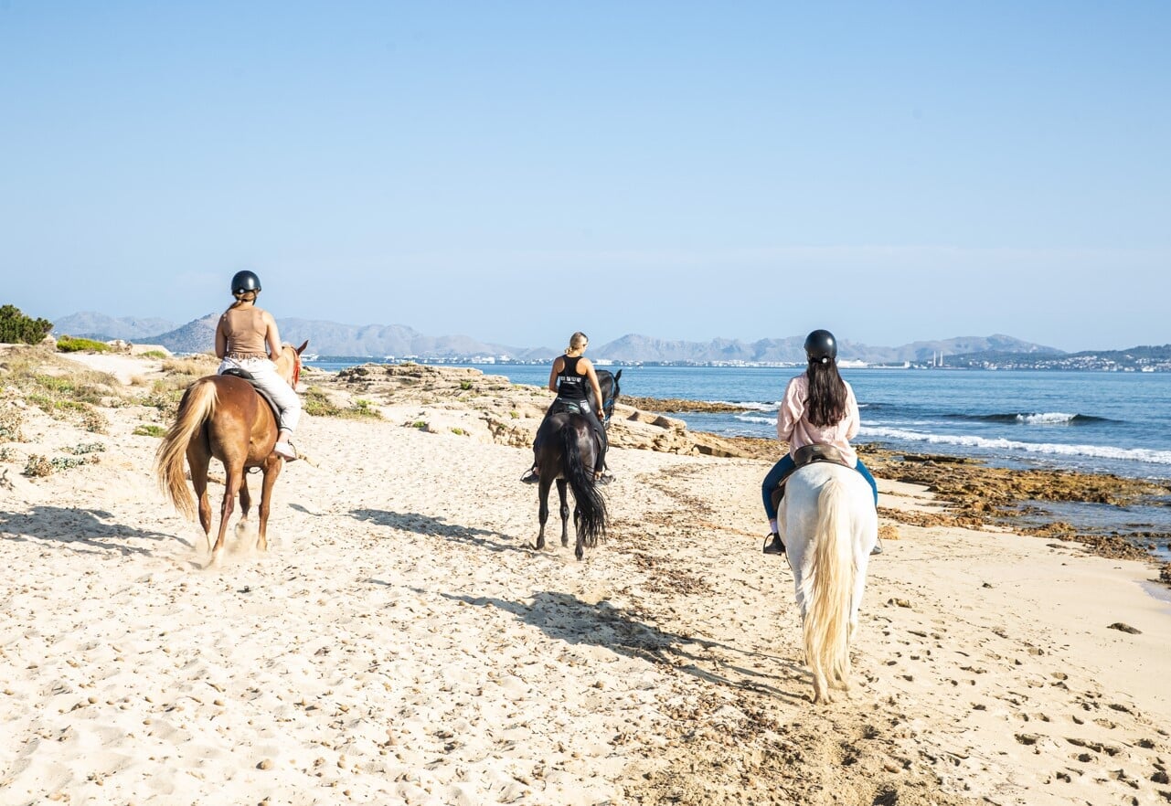 Three people riding horses on the beach by the sea with a black, brown and white horse during a horseback excursion in Mallorca